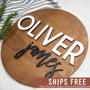 May include: A circular wooden sign with a smooth, medium-brown stained finish. The name OLIVER is displayed in large, white, uppercase sans-serif letters, while the name james is written below in smaller, black, lowercase script lettering.