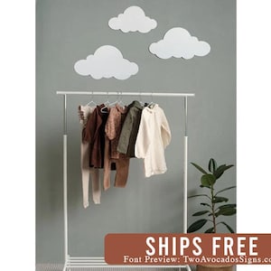 May include: Three white cloud-shaped wall decorations mounted on a gray wall above a white metal clothing rack. The rack holds several garments in neutral tones, including brown, olive green, and cream-colored shirts and pants on hangers.