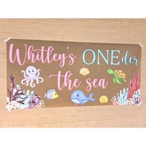 May include: A brown banner with the text "Whitley's ONE der the sea" in pink and blue. The banner features illustrations of an octopus, fish, whale, turtle, seashells, and coral.