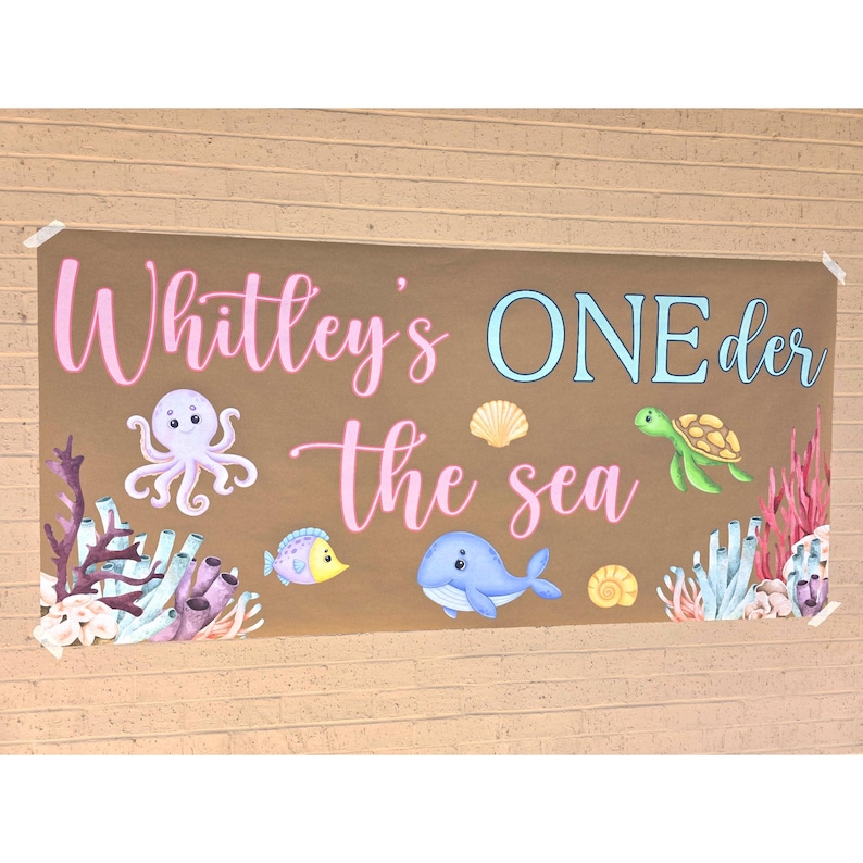 May include: A brown banner with the text "Whitley's ONE der the sea" in pink and blue. The banner features illustrations of an octopus, fish, whale, turtle, seashells, and coral.