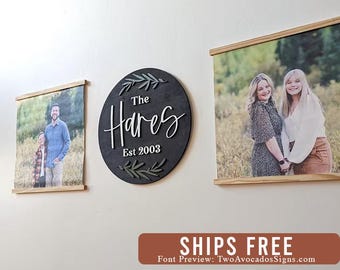 May include: Three wall hangings. Two rectangular photo prints with wooden dowels at the top and bottom. A round black sign with white script text that reads "The Hares Est 2003" and green leaf accents. The photos show people.