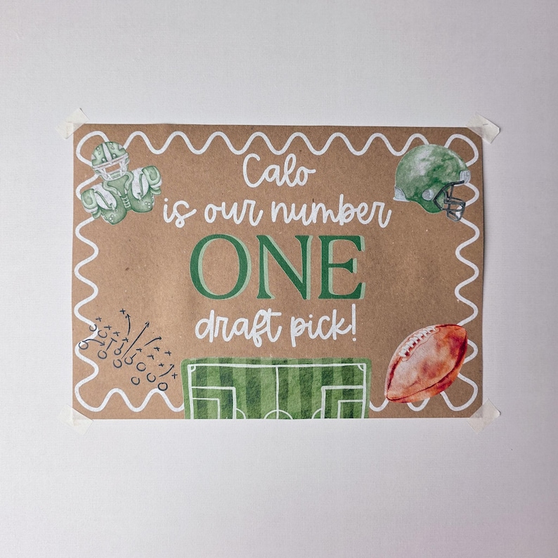 May include: A rectangular brown paper sign with a white scalloped border featuring the text Calo is our number ONE draft pick! The design includes illustrations of a green football helmet, shoulder pads, a football, and a field diagram.