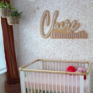 May include: A white crib with a wooden top rail and a pink mattress. The crib is in a nursery with a white floral wallpaper and a wooden name sign that reads "Claire Elizabeth".