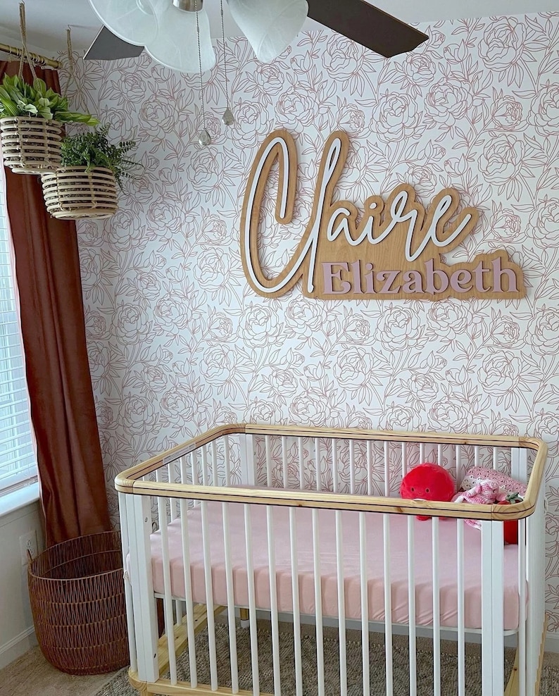 May include: A white crib with a wooden top rail and a pink mattress. The crib is in a nursery with a white floral wallpaper and a wooden name sign that reads "Claire Elizabeth".