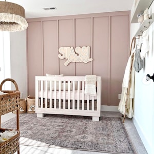 K&ouml;nnte beinhalten: Ein wei&szlig;es Babybett mit einer rosa-wei&szlig;en Decke in einem Kinderzimmer mit einer rosafarbenen Wand, einem gewebten Teppich und einem Holzschild mit der Aufschrift "Stassi James".