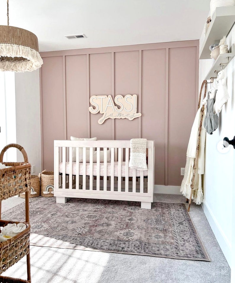 K&ouml;nnte beinhalten: Ein wei&szlig;es Babybett mit einer rosa-wei&szlig;en Decke in einem Kinderzimmer mit einer rosafarbenen Wand, einem gewebten Teppich und einem Holzschild mit der Aufschrift "Stassi James".