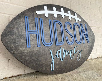 May include: A wooden football-shaped sign with the name "HUDSON james" in blue and light blue lettering.