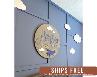 Puede incluir: Un letrero de pared circular de madera con el nombre Henry Alexander en letras azules cursivas y de molde, acompañado de un icono de avión azul y varios recortes de madera blanca en forma de nube dispuestos alrededor.