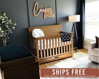 May include: A wooden cot with vertical slats and a bottom storage drawer, featuring a personalised wooden wall sign above it that reads Cooper Jacob in a script and block font combination, set against a dark grey wall in a nursery room.
