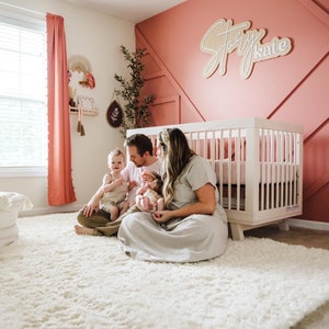 K&ouml;nnte beinhalten: Eine vierk&ouml;pfige Familie sitzt auf einem wei&szlig;en, flauschigen Teppich in einem Kinderzimmer. Die Eltern halten ein Baby und ein Kleinkind. Das Kinderzimmer hat ein wei&szlig;es Babybett mit einer rosa Decke, eine rosa Wand mit einem Holzschild, auf dem "Story Kate" steht, und ein Fenster mit rosa Vorh&auml;ngen.