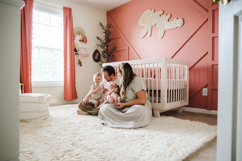K&ouml;nnte beinhalten: Eine vierk&ouml;pfige Familie sitzt auf einem wei&szlig;en, flauschigen Teppich in einem Kinderzimmer. Die Eltern halten ein Baby und ein Kleinkind. Das Kinderzimmer hat ein wei&szlig;es Babybett mit einer rosa Decke, eine rosa Wand mit einem Holzschild, auf dem "Story Kate" steht, und ein Fenster mit rosa Vorh&auml;ngen.