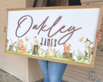 Op de afbeelding: Een houten bord met de namen "Oakley James" in een cursief schrift. Het bord is versierd met een boslandschap met een vos, een uil, een eekhoorn en een hert.