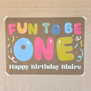 May include: A brown paper banner with the words "FUN TO BE ONE" in colorful bubble letters. Below, it reads "Happy Birthday Blaire" in white. The banner is taped to a brick wall.