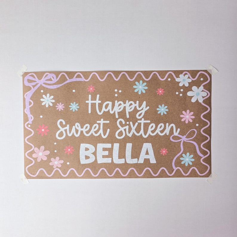 May include: A rectangular brown sign featuring white text that reads Happy Sweet Sixteen BELLA. The border is decorated with a light pink wavy line, bows in the corners, and small light blue and pink flower illustrations scattered throughout.