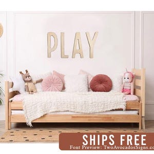 Pu&ograve; includere: Un cartello in legno PLAY in un tono di legno chiaro, montato su una parete bianca sopra un divano letto in legno con biancheria da letto bianca e cuscini rosa e bianchi. Il divano letto &egrave; decorato con animali di peluche.