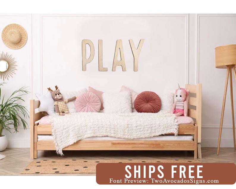 Pu&ograve; includere: Un cartello in legno PLAY in un tono di legno chiaro, montato su una parete bianca sopra un divano letto in legno con biancheria da letto bianca e cuscini rosa e bianchi. Il divano letto &egrave; decorato con animali di peluche.