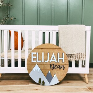 Op de afbeelding: Een rond houten bord met een bergketenontwerp en de namen Elijah Dean in witte letters. Het bord hangt aan een wit babybedje met een beige deken en een bruin kussen.
