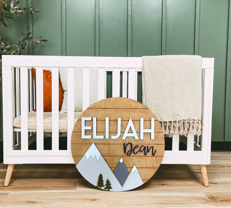 Op de afbeelding: Een rond houten bord met een bergketenontwerp en de namen Elijah Dean in witte letters. Het bord hangt aan een wit babybedje met een beige deken en een bruin kussen.