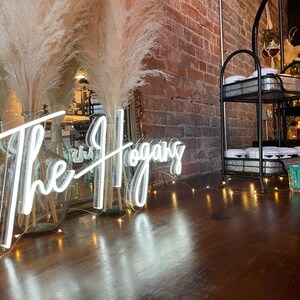 K&ouml;nnte beinhalten: Ein wei&szlig;es Neon-Schild mit dem Text "The Hogans" auf einer Holzoberfl&auml;che mit einer Lichterkette. Zwei hohe Glasvasen mit getrocknetem Pampasgras stehen vor dem Schild.