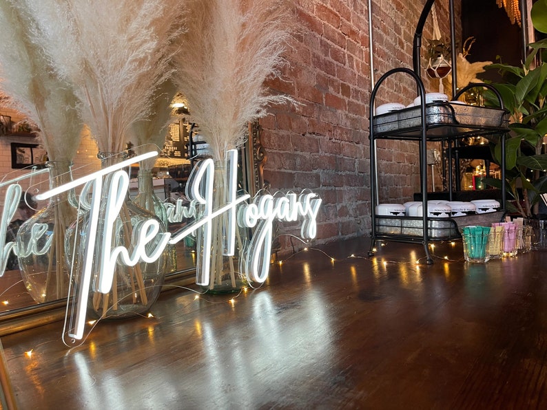 K&ouml;nnte beinhalten: Ein wei&szlig;es Neon-Schild mit dem Text "The Hogans" auf einer Holzoberfl&auml;che mit einer Lichterkette. Zwei hohe Glasvasen mit getrocknetem Pampasgras stehen vor dem Schild.