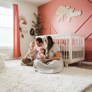 May include: A family of four sits on a white shag rug in a nursery. The parents are holding their two children, a baby and a toddler. The nursery has a white crib with a pink and white bedding set. The wall behind the crib is painted a light pink color and has a wooden sign that says "Story Kate".