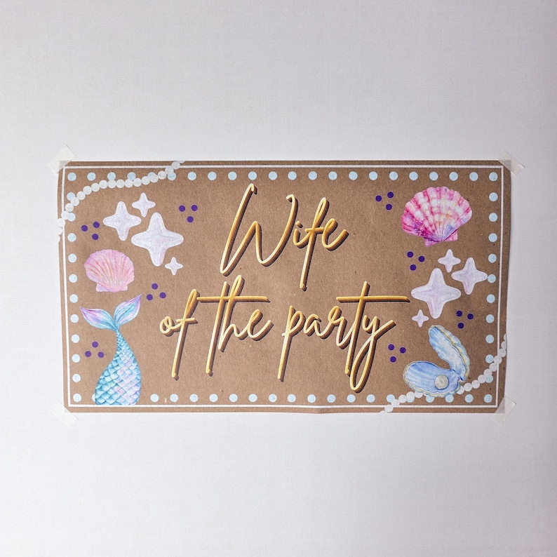 May include: A rectangular sign with the words "Wife of the party" in gold script. The sign is brown with white pearl and dot borders, and features illustrations of seashells, starfish, and a mermaid tail.