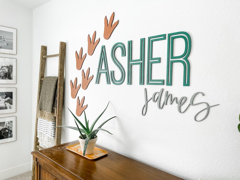 Pu&ograve; includere: Una cameretta con una scala di legno, una coperta e una pianta. Il muro ha un cartello con il nome verde e bianco che dice "ASHER JAMES" con impronte di dinosauro marroni intorno.