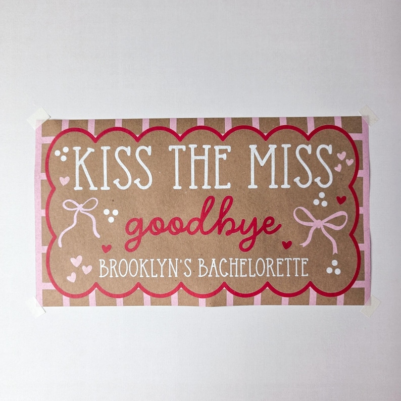May include: A rectangular sign with the text "KISS THE MISS goodbye" in white and red lettering. The sign is brown with a pink scalloped border and pink accents, including hearts and bows. The text "BROOKLYN'S BACHELORETTE" is also included.