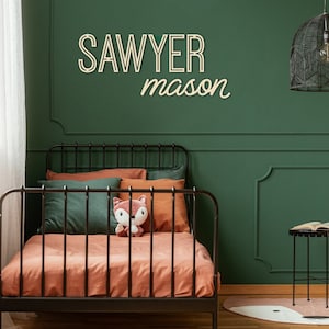 Pode incluir: Uma parede verde com uma estrutura de cama de metal preto e um conjunto de roupa de cama marrom e verde. A parede tem uma placa de madeira com o nome "Sawyer mason". Um lustre preto em gaiola pendura do teto.