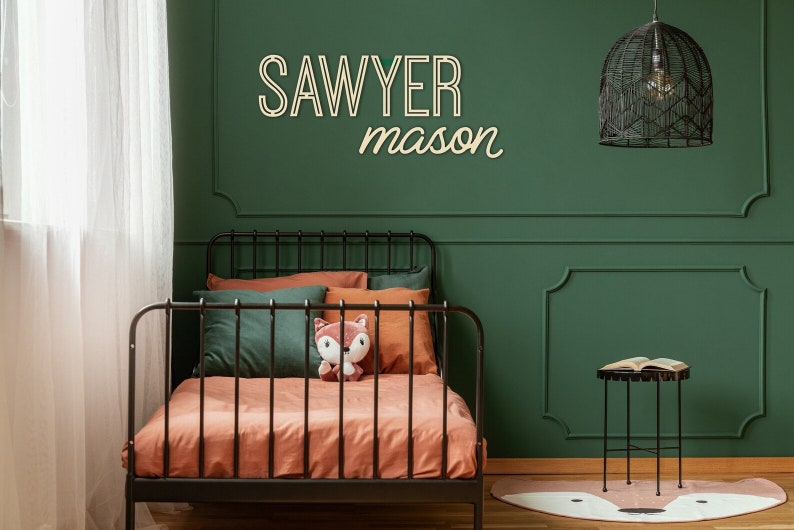 Pode incluir: Uma parede verde com uma estrutura de cama de metal preto e um conjunto de roupa de cama marrom e verde. A parede tem uma placa de madeira com o nome "Sawyer mason". Um lustre preto em gaiola pendura do teto.