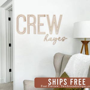 May include: Wooden wall decor with the word "CREW" and the name "hayes" in a cursive font. The decor is mounted on a white wall in a room with a white couch and a lamp.