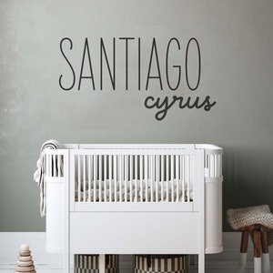 Pu&ograve; includere: Un lettino bianco con un materasso bianco e una coperta bianca. Il lettino &egrave; in una cameretta con una parete grigia e uno sgabello di legno con un cuscino bianco. La parete ha un adesivo in vinile nero con i nomi "Santiago Cyrus".