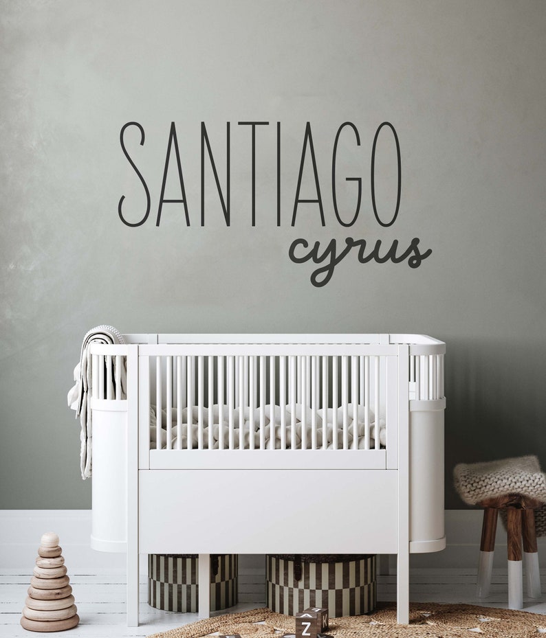 Pu&ograve; includere: Un lettino bianco con un materasso bianco e una coperta bianca. Il lettino &egrave; in una cameretta con una parete grigia e uno sgabello di legno con un cuscino bianco. La parete ha un adesivo in vinile nero con i nomi "Santiago Cyrus".