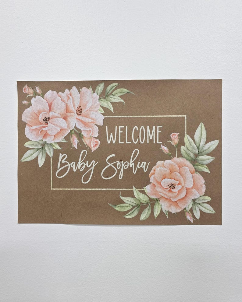 May include: A brown card with the words "WELCOME Baby Sophia" in white script, framed by a white rectangle. The card is decorated with watercolor-style peach-colored roses and green leaves.