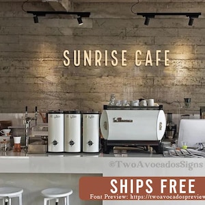 May include: Sunrise Cafe sign with a white background and a brick wall behind it. The sign is made of metal and has a modern font. There are coffee machines and other equipment in the background.