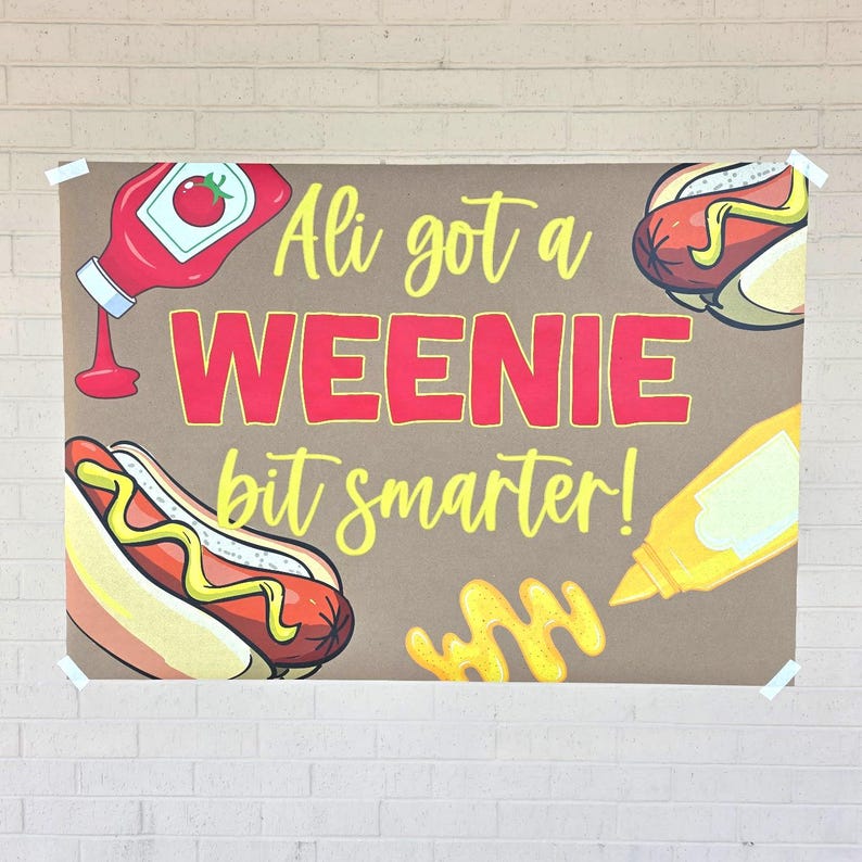 May include: A brown poster with the text "Ali got a WEENIE bit smarter!" in red and yellow. The poster features illustrations of hot dogs, ketchup, and mustard.