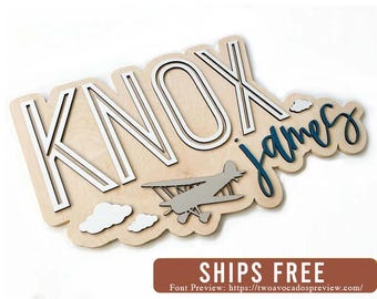 May include: A custom wooden wall sign featuring the name KNOX in large white block letters and the name james in dark blue cursive script. A gray biplane silhouette and white cloud cutouts are layered onto the light wood backing board.
