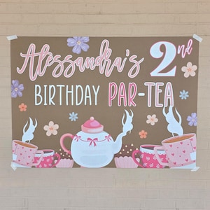 May include: A brown poster with the text "Alessandra's 2nd BIRTHDAY PAR-TEA" in pink and white. The poster features illustrations of teacups, a teapot, and flowers in pink, white, and peach colors. The poster is taped to a brick wall.