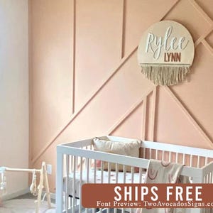 May include: A circular wooden wall sign featuring the name Rylee in white script and the name LYNN in brown block letters, finished with a beige macrame fringe at the bottom. The sign is mounted on a peach-colored wall with geometric trim.