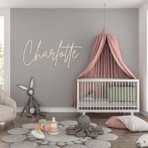 May include: A nursery room featuring a white crib with a dusty pink canopy, a wooden wall sign reading Charlotte in cursive script, a gray rabbit plush, a gray octopus plush, a pastel stacking ring toy, and a gray circular petal-patterned rug.