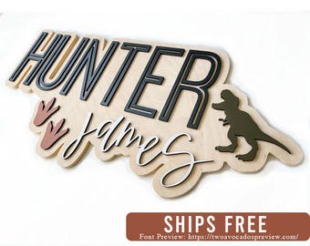 May include: A custom wooden wall sign featuring the name Hunter James in layered lettering. The name Hunter is in bold black block letters, while James is in white cursive script. A dark green dinosaur silhouette and two light brown dinosaur footprints are attached.