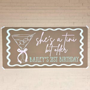 May include: A brown banner with a light blue wavy border and the text "she's a tini bit older" in pink script. A martini glass illustration is on the left. The text "BAILEY'S 21ST BIRTHDAY" is at the bottom.