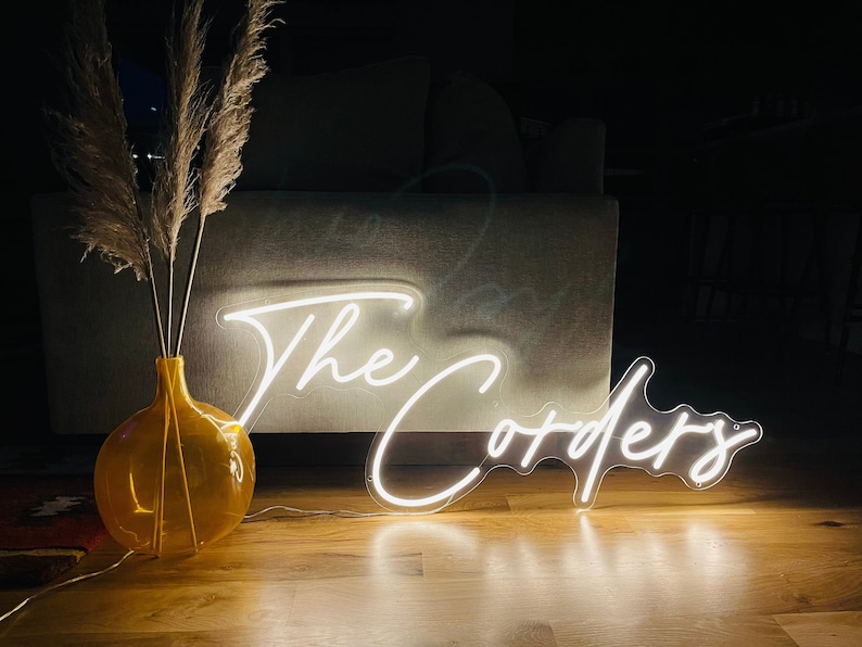 K&ouml;nnte beinhalten: Ein wei&szlig;es Neon-Schild mit dem Text "The Corders" in einer kursiv geschriebenen Schrift. Das Schild leuchtet und steht auf einem Holzboden. Im Vordergrund steht eine Vase mit getrocknetem Pampasgras.