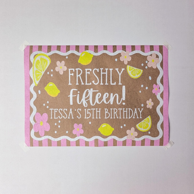 K&ouml;nnte beinhalten: Ein rechteckiges Schild mit brauner Mitte und einem rosa-wei&szlig; gestreiften Rand. Der Text lautet FRESHLY fifteen! TESSA'S 15TH BIRTHDAY. Das Design zeigt gelbe Zitronenscheiben, ganze gelbe Zitronen sowie kleine rosa und wei&szlig;e Blumen.