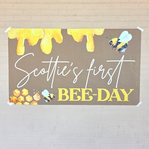 May include: A brown banner with yellow honey drips at the top and the text "Scottie's first BEE-DAY" in white and yellow. Watercolor bees and honeycomb accents decorate the banner, perfect for a first birthday celebration.