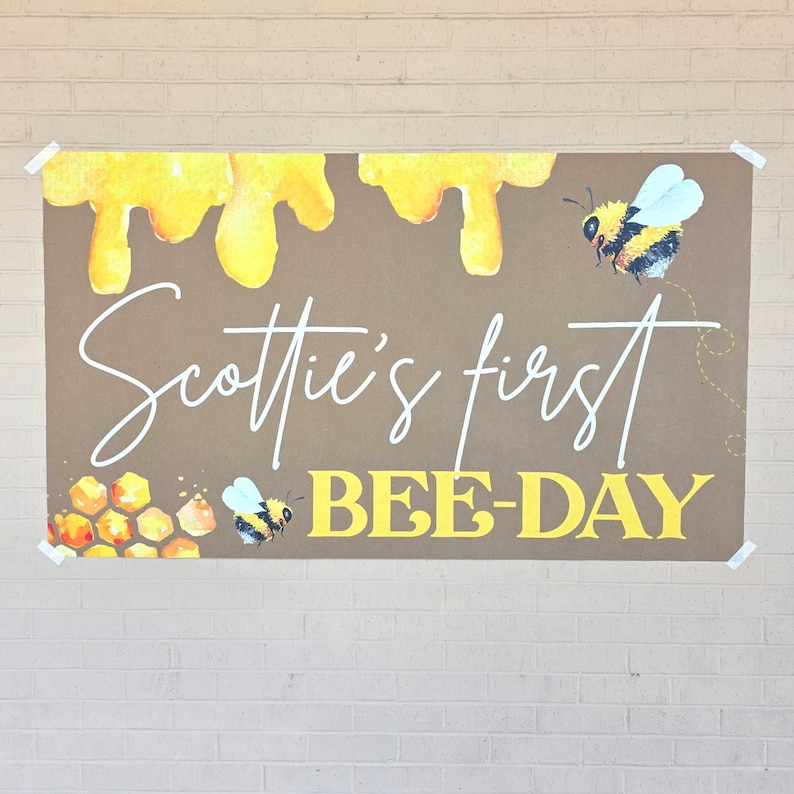 May include: A brown banner with yellow honey drips at the top and the text "Scottie's first BEE-DAY" in white and yellow. Watercolor bees and honeycomb accents decorate the banner, perfect for a first birthday celebration.