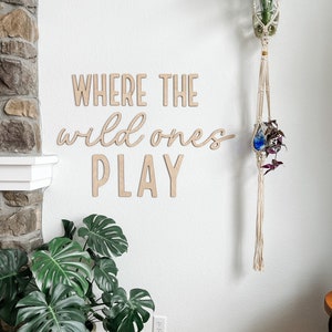 May include: Light-colored wooden wall lettering spelling out the phrase WHERE THE wild ones PLAY in a combination of block and cursive fonts. The letters are mounted on a plain white wall, positioned between a large green plant and a macrame hanger.