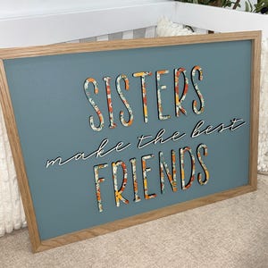 May include: A teal blue wooden sign with a wood frame. The sign says "Sisters make the best friends" in white lettering with a floral pattern.