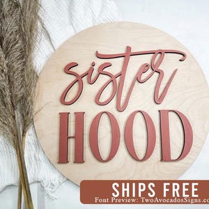 May include: A circular light-colored wooden sign featuring the word sister in a dark pink cursive script font positioned above the word HOOD in a dark pink block letter font. The sign has a smooth finish and a natural wood grain texture.