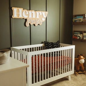 May include: A white crib with a red and white patterned sheet and a black and white pillow. The crib is in a room with a green wall and a wooden shelf with books. A wooden name sign with the names "Henry James" is on the wall above the crib.